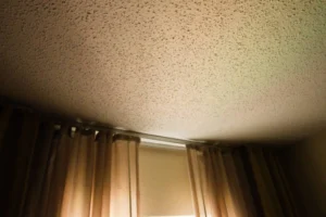 A ceiling with a popcorn texture and a window with curtains below it.